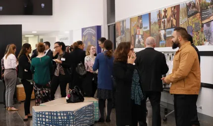 Group of Indigenous people, young and old, talk amongst themselves in a room with art on the walls, a white ceiling and dark tiled floor.