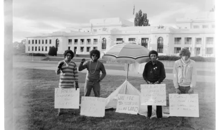 Four men in casual wear stand on grass and either side of a beach umbrella. In front of them are small hand written signs. In the background is a road and then a white building with many windows.