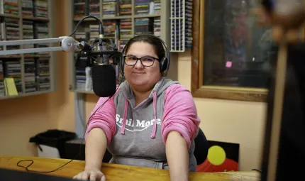 Aboriginal woman wearing glasses and a grey and pink top sits at a desk in front of a microphone. In the background is a rack of discs, a window and an Aboriginal flag.