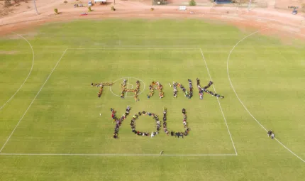 A bird’s eye view of people on a green oval. They stand close together and form the words ‘Thank you’. In the background is a brown road, buildings and more people.