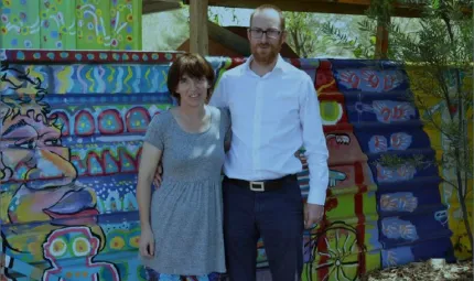 A woman in a grey dress and colourful tights and a man in white shirt and blue trousers stand in front of a painted fence featuring a face, a bicycle and other designs of blue, green, red, yellow and purple.