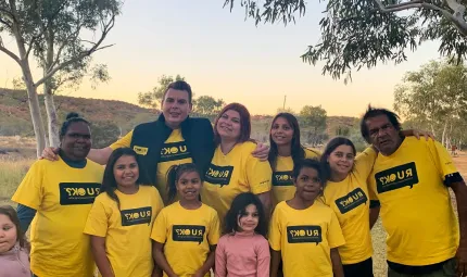 Group of Indigenous adults, youth and children in yellow t-shirts with the letters RUOK on them. Two young girls are dressed in pink tops. In the background is a small hill and trees.