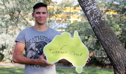 Young Indigenous man wearing grey t-shirt standing beneath a tree and holding a cardboard cut-out of Australia featuring a dot with an arrow to the words Tennant Creek.