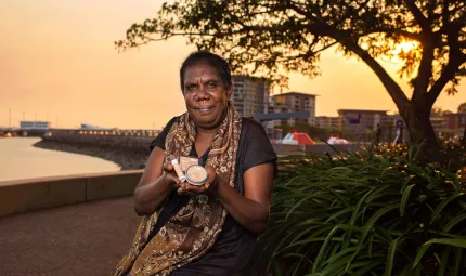 An Aboriginal woman in dark dress and patterned scarf sits next to a bush and holds three small containers in her hands. In the background is water, a wharf, a tree and buildings.