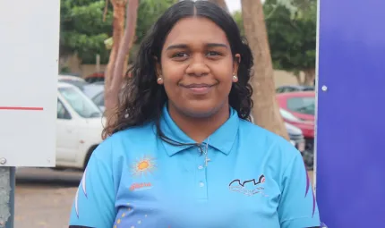 A young woman with long black hair wearing a light blue polo shirt stands in front of trees and some parked cars.