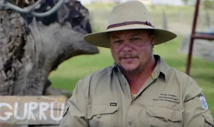 An Aboriginal man wears a broad brimmed hat and a khaki uniform. In the background is a tree, grass and metal structure.