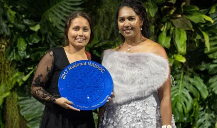 Two young Torres Strait Islander women standing with a background of plants. The young woman on the left is holding a large blue plate.