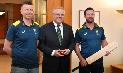 Three men stand in a room. In the background is a white wall and a doorway through to neighbouring room. The man on the left wears a blue polo shirt and blue jeans. The man in the middle wears a dark suit. The man on the right wears darks trousers.