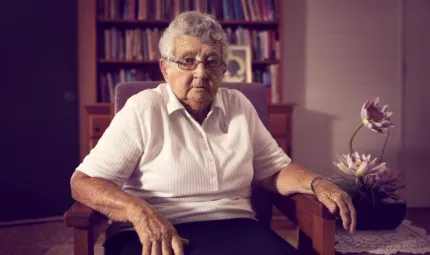 Photograph of an elderly woman wearing glasses and a white top, she is sitting on a wooden dining chair. Behind her is a bookshelf, and next to her is a plant.