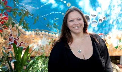 Indigenous woman dressed in black stands in front of colourful background made up of blue, orange and green layers featuring red and white flowers, green leaves and Aborginal designs.