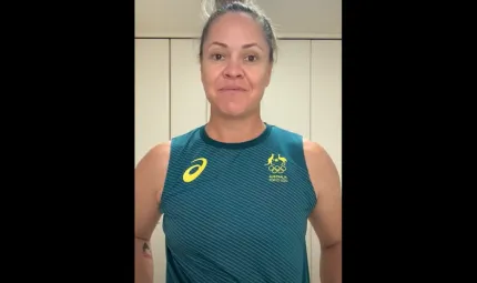 A woman stands in front of a cream wall. She is wearing a green and yellow Olympic singlet.