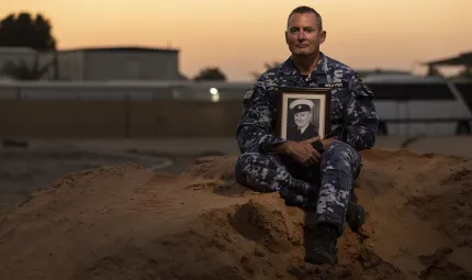 A man dressed in an air force uniform and holding a photo of another man in uniform sits on a dirt mound with a setting sun and buildings in the background.