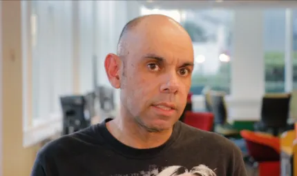 Balding man in black t-shirt in foreground with office furniture and windows in the background.