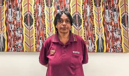 Aboriginal woman in maroon polo shirt stands in front of an Aboriginal painting of featuring vertical patterns of lines and animals and spears and other figures.