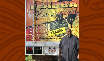 Aboriginal man in grey shirt stands in front of the rear door of a truck. The door is painted with red, yellow and black designs and includes the following words: TEABBA Media, TEABBA N.T. Outback Australia, www.teabba.com