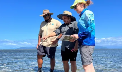 Three people stand in sallow sea water under which is sea grass. They are dressed in work wear and wear hats. In the background is an island with clouds and blue sky beyond that.