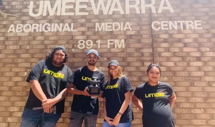 Four Aboriginal young adults (2 men and 2 women) in black t-shirts with UMEEtv printed on them stand in front of a brick wall. On the wall are the words: Umeewarra Aboriginal Media Centre 89.1 FM