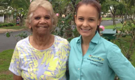 Elderly Indigenous woman in yellow and grey dress stands with arm around young Indigenous woman dressed in aqua coloured shirt in front of a flowering bush in a suburban street.