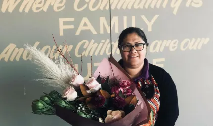 Aboriginal woman in black top holds flowers in front of a sign which says ‘Where Community is, Family And our Children are our Future’.