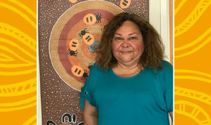 Indigenous Primary School teacher standing front of Indigenous artwork of Honey ants and dots in circles on a brown background.