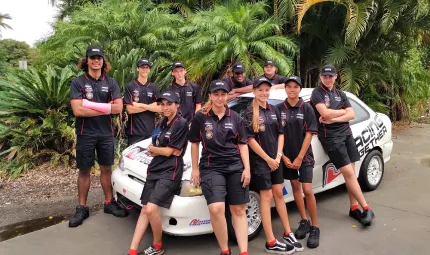 Group of young Indigenous Australians sit on, or stand behind a white racing car. They each wear a blue uniform of polo shirt, shorts and cap. In the background are ferns and trees.