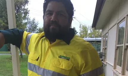 An Aboriginal man with beard and wearing a yellow safety shirt stands next to a beige coloured building. In the background is green grass, trees and another building.