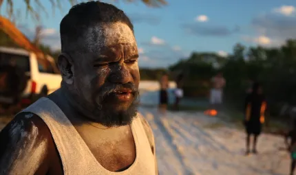 Aboriginal man Timmy Burarrwanga in foreground with others in background outdoors, standing on sandy soil.