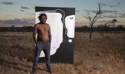 A man wearing dark pants and without a top stands on semi-arid and grassy soil. Behind him is a large black and white painting. In the background are a few trees, a flat landscape and darkening sky.