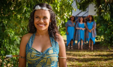 A bride is in traditional Tiwi wedding dress, of bright blue and lime green and is smiling towards the camera. She is outside on a sunny day and there are 3 bridesmaids in blue dresses in the background.