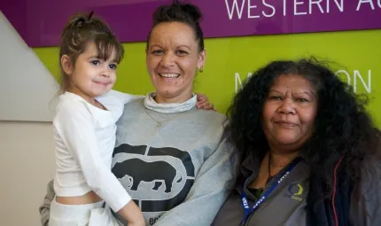 Two Indigenous adult women, one dressed in light grey shirt and holding a child dressed in white clothes, the other in a dark grey shirt.