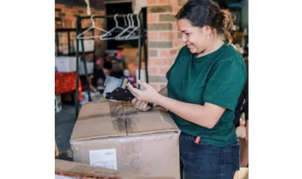 A young woman in a green top and blue jeans applies sticky tape to a large cardboard box. She works in a room with brick walls which contains different storage objects.