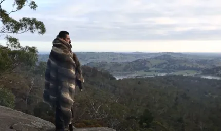 A young adult man wrapped in animal skins stands on a rocky outcrop and looks over tree covered rolling hills below.