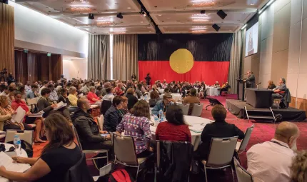 Large gathering of people in a hall, one standing at a rostrum and most seated at tables or on seat rows. The carpet is red, the walls are beige and the curtains are brown. Covering the far end wall is the Aboriginal flag.