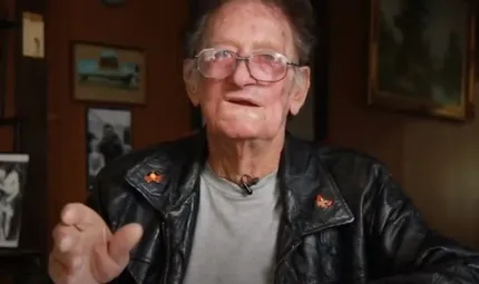 A mature man in leather jacket and wearing glasses sits in a room. In the background is a wall covered in photos and other images.