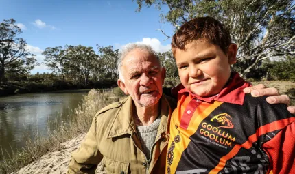 An Aboriginal man in brown jacket and boy in red, black and yellow shirt, stand near a river behind them. Also in the background are trees, grass and a blue sky.