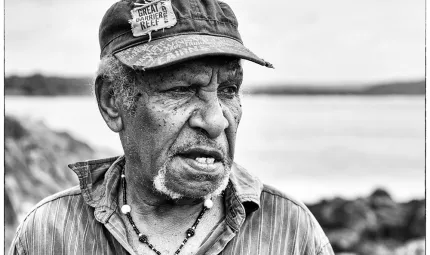 An elderly man in a baseball cap and lined shirt looks to left of camera. In the background are rocks and water.