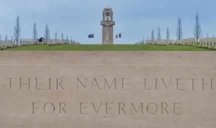 Tall tower flanked by Australian and French flags and to left and right are cemetery headstones with trees in the background. In the foreground is a carved stone with the words Their Name Liveth For Evermore