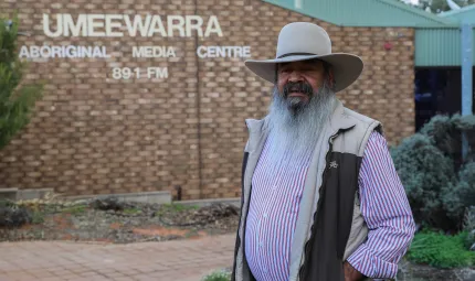 Aboriginal man with long white beard wearing a hat, striped shirt and vest stands in front of a brick building with the following words on the wall: Umeewarra Aboriginal Media Centre 89.1 FM.