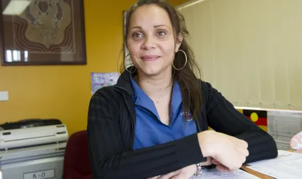 Woman dressed in black cardigan and blue shirt in an office and sitting at a desk covered with papers.