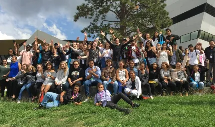 Large group of Indigenous young adults and their teachers sitting and standing on a lawn outside a large building and in front of a tree.