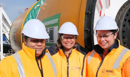 Three smiling Indigenous women in yellow jackets and white helmets stand in front of a large yellow, green and white machine.