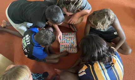 The arrival of the Warlpiri Encyclopaedic Dictionary in Yuendumu, Northern Territory. Five children and an adult are bent over the Warlpiri Encyclopaedic Dictionary. The cover of the dictionary has photos of people who have contributed to it.