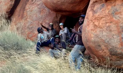 Group of people sitting under large rock formation. In the foreground is long grass.