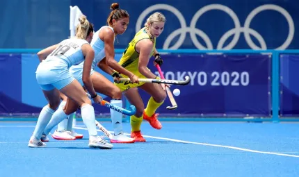 Three women, two dressed in blue uniforms and the other in yellow, hold hockey sticks and chase a small white ball on a blue surface. In the background are the words Tokyo 2020 and 5 rings overlaid in a line.