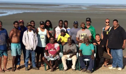 A group of Indigenous men stand or sit on chairs in front of a mud flat and estuary in the background.