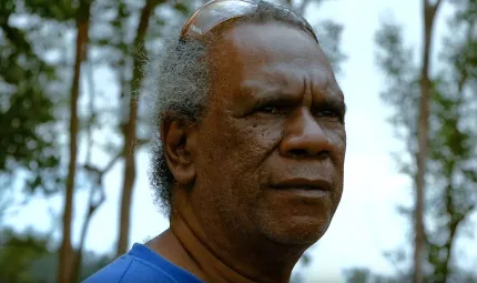 A head photo of an Indigenous man in blue shirt with sunglasses on his head. In the background are trees and sky.