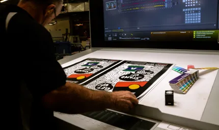 A man stands at a drafting table on which are two mainly black and white posters. To the right of the posters is a colour matching gauge. In the background is a large video screen.