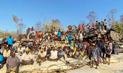 A large group of men, women and children wearing casual or work wear sit or stand on a rocky outcrop facing the camera. In the background are a few trees and a blue sky.