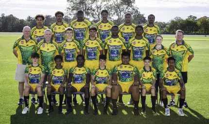 A group of mainly young men in sporting attire sit and stand on a grassed playing field in three rows. They each wear a blue, yellow and green jersey. In the background is green grass and trees.