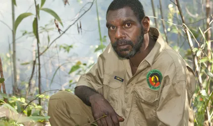 Indigenous man dressed in a ranger uniform sits in foreground with foliage in the background.